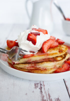 Fork cutting into a stack of three Strawberry Buttermilk Pancakes with Fresh Strawberry Syrup topped with sliced strawberries and whipping cream.