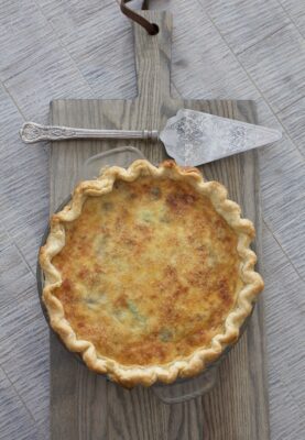 ABK's Basic Favorite Quiche on a cutting board on a grey tabletop next to a silver serving spoon.
