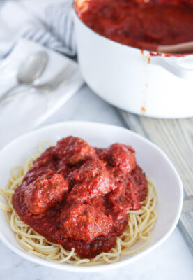 Close up of white bowl of spaghetti and meatballs. White pot of sauce in background.
