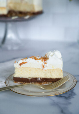 Slice of Better than Cheesecake Factory Coconut Cheesecake on a china plate with gold fork. Cheesecake on stand in the background.