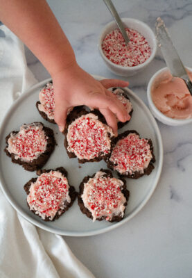 Chocolate Cookie with Cream Cheese Frosting and Peppermint on a cookie sheet with bowls of frosting and peppermint bits. Small child's hand reaching for a cookie.